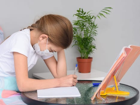 A girl in a medical mask does homework at a table in front of the bed