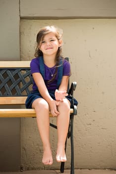 A little girl sits on a bench outside in the back garden of her home.