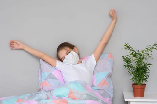 A recovering girl in a medical mask woke up in the morning and stretches herself in bed
