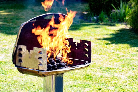 Close-up of the frozen moment of the blazing flames in a charcoal grill with green grass background of a garden. Prepearing of a barbecue party at a sunny spring day. BBQ concept. Macro.