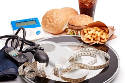 Fast food and medical devices tonometer (blood pressure measuring device) and blood glucose meter, scales and measuring tape on a white background. Concept of unhealthy diet and health.
