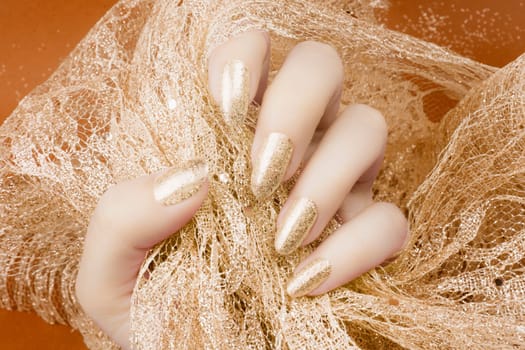 Female hand with glittered gold nails manicure is holding gold decoration on orange background.