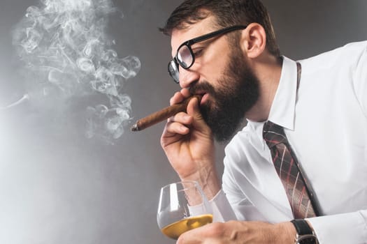 The elegant bearded man is smoking cigar and drinking alcohol on gray background.