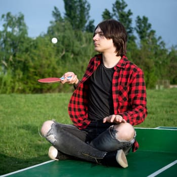 The teen boy or young guy is sitting on table tennis table and playing with racket and ball.
