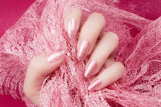 Female hand with glittered rose nails manicure is holding rose decoration on pink background.