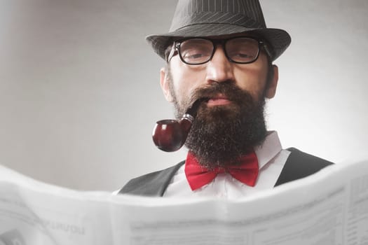 The elegant old-fashioned bearded hipster middle aged man with tobacco pipe is reading newspaper.