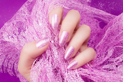 Female hand with glittered pink nails manicure is holding pink decoration on pink background.