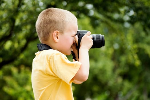 The little blond hair boy photographer is photographing outdoors on green background.