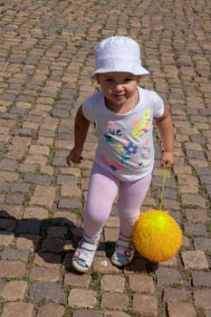 Adorable little happy baby girl walking in a pedestrian street with a large yellow ball  on a sunny summer day.