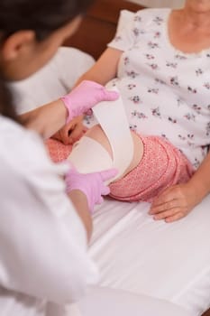Female doctor bandages senior woman leg at home or in nursing home. Medicine, age, support, health care and people concept - doctor caring senior patient.  Focus on nurse hand.