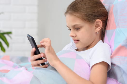 The girl lies in bed and plays in a mobile smartphone