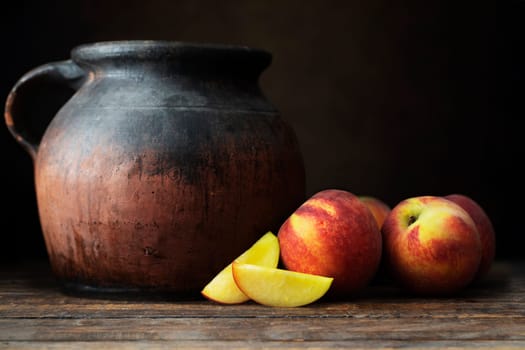 Fresh nectarines, whole and sliced, next to a rustic pottery jug, on  awooden table with a dark background