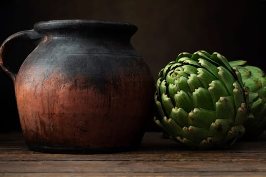 Two fresh artichokes and a rustic pottery jug on a dark wood surface.