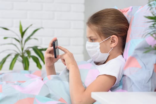 The girl lies in bed in a medical mask and does web surfing on the phone