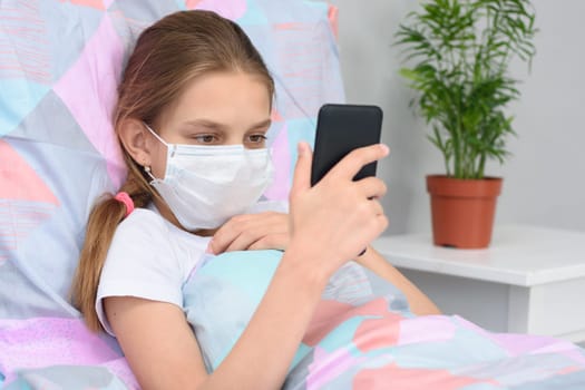 A girl in a medical mask lies in bed and looks at the phone screen