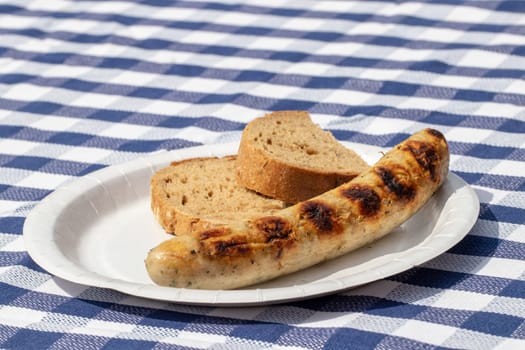 Closeup of a roasted sausage with bread on paper plate on blue checkered table cloth. Macro photograph.