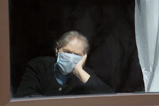 An old grandmother in a protective mask looks out the window at self-isolation. Elderly woman at quarantine at home.