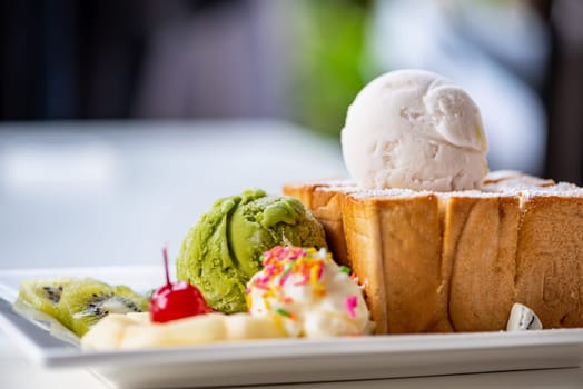 Closeup Honey Toast bread served with green tea and coconut milk ice cream scoop, decorate with cherry, apple, kiwi fruit, and cream sugar topping sprinkle on a white dish sweet cool snack in summer