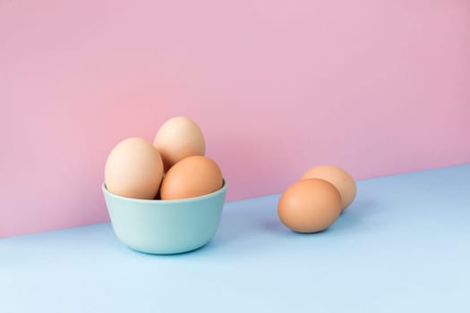 Minimalist image of several brown eggs in a bowl on a light blue and pink background. Copy space available.