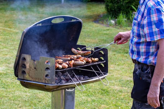 A man at the charcoal grill is grilling steaks, grill sausages and chicken wings and using barbecue tongs. Selective focus on the grill against a green lawn background on a sunny spring day. BBQ concept. Macro.