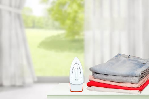 Household laundry ironing. Close-up of a red electrical iron and a stack of ironed clothes on white board over blurred curtains background. Advertising laundry service.
