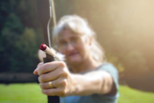Woman with gray hair aiming with bow and arrow in nature and sun. Concept for the fight to reach the target