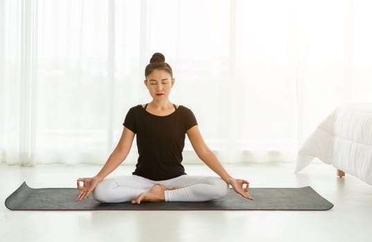 Middle aged women practicing yoga in Easy seat pose or Sukhasana with with mudra. Meditation with yoga in white bedroom after wake up in the morning. Concept of exercise, relaxation and healthcare.
