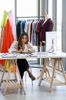 Young adult asian fashion designer drawing and sketching her work at home as atelier studio with colour palette. Using for entrepreneur small business startup concept.
