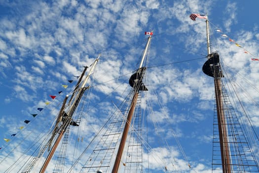 The tree masts of a large tall ship with there flags under a sonny sky.with some white clouds.