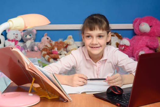 The girl in the children's room sits at the desk with a smile and does her homework