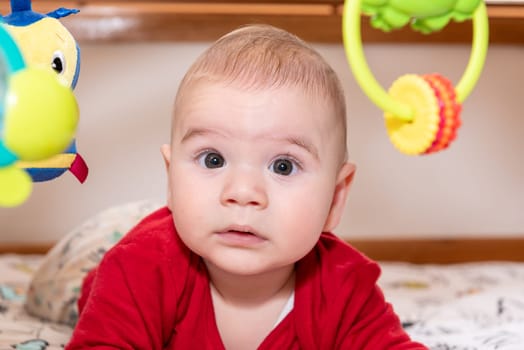 Cute little baby boy during tummy time looking at camera, 6 months old child with curiosity expression on his face surrounded by colourful toys.