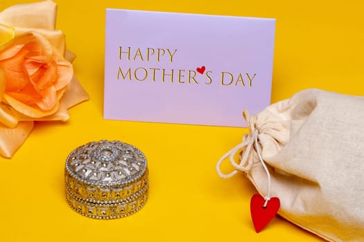 Mothers day greeting card. Close-up of a Jewellery box and beautiful yellow blossom of flower and a tag with inscription Happy mothers day and fabric gift drawstring bag on yellow background. Beauty for the mom. Macro.