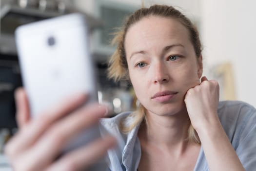 Worried woman indoors at home kitchen using social media apps on phone for video chatting and stying connected with her loved ones. Stay at home, social distancing lifestyle.
