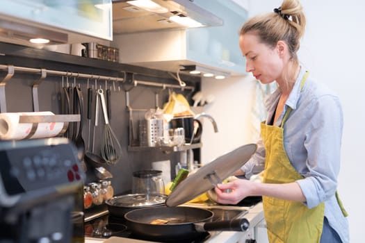 Stay at home housewife woman cooking in kitchen, stir frying dish in a saucepan, preparing food for family dinner. Blonde caucasian model in her 30is, real cooking not looking in camera.