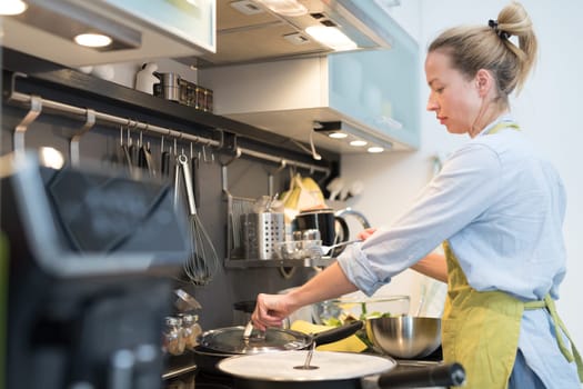 Stay at home housewife woman cooking in kitchen, stir frying dish in a saucepan, preparing food for family dinner. Blonde caucasian model in her 30is, real cooking not looking in camera.