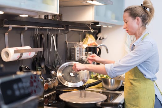 Stay at home housewife woman cooking in kitchen, stir frying dish in a saucepan, preparing food for family dinner. Blonde caucasian model in her 30is, real cooking not looking in camera.