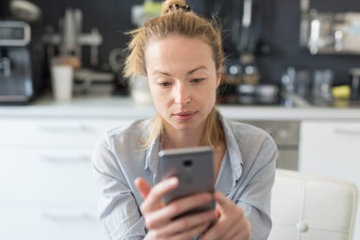 Worried woman indoors at home kitchen using social media apps on phone for video chatting and stying connected with her loved ones. Stay at home, social distancing lifestyle.