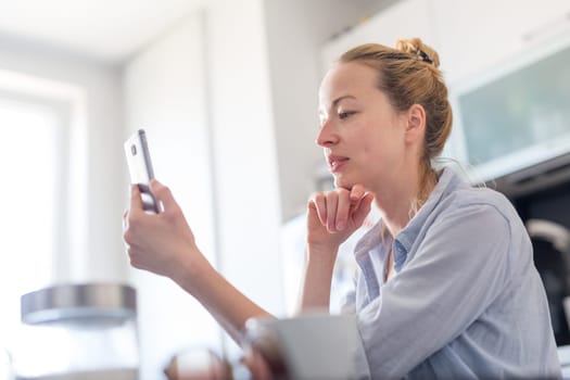 Young smiling cheerful woman indoors at home kitchen using social media on phone for video chatting and stying connected with her loved ones. Stay at home, social distancing lifestyle.