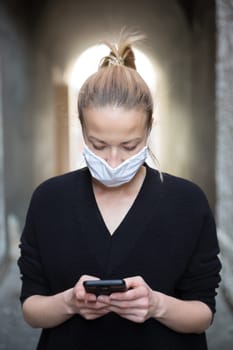 COVID-19 pandemic coronavirus. Casual caucasian woman at medieval city street using mobile phone, wearing protective face mask against spreading of coronavirus and disease transmission.