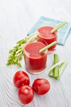 Two glasses with fresh tomato juice, celery, parsley and ripe tomatoes on light wooden background. Top view