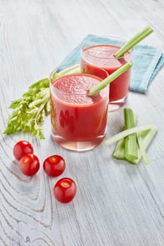 Two glasses with fresh tomato juice, celery, parsley and ripe tomatoes on light wooden background. Top view