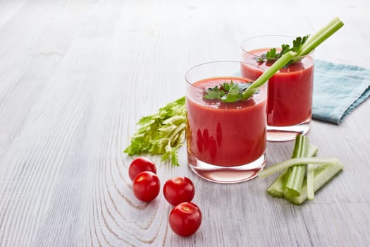Two glasses with fresh tomato juice, celery, parsley and ripe tomatoes on light wooden background. Top view