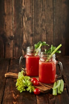 Two glasses with fresh tomato juice, celery, parsley and ripe tomatoes on dark brown wooden background. Healthy beverage, vegetable juice