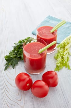 Two glasses with fresh tomato juice, celery, parsley and ripe tomatoes on light wooden background. Top view