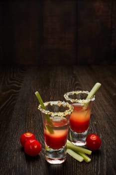 Two shot glasses with cocktail Bloody Mary, celery and ripe tomatoes on dark wooden background