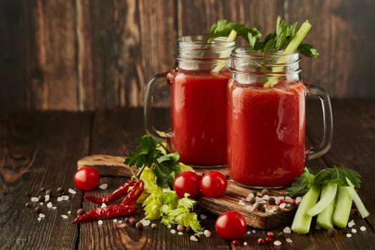 Two glasses with fresh tomato juice, celery, parsley, spices and ripe tomatoes on dark brown wooden background with copy space. Healthy beverage, vegetable juice