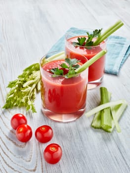 Two glasses with fresh tomato juice, celery, parsley and ripe tomatoes on light wooden background. Top view