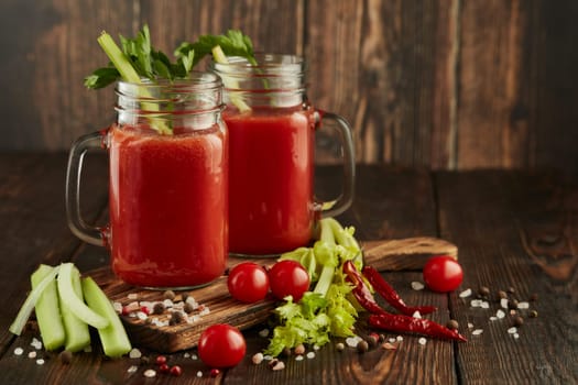 Two glasses with fresh tomato juice, celery, parsley, spices and ripe tomatoes on dark brown wooden background with copy space. Healthy beverage, vegetable juice