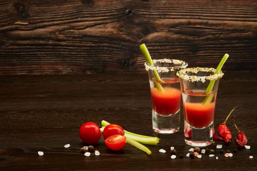 Two shot glasses with cocktail Bloody Mary, celery, spices and ripe tomatoes on dark wooden background with copy space