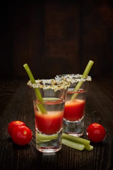 Two shot glasses with cocktail Bloody Mary, celery and ripe tomatoes on dark wooden background